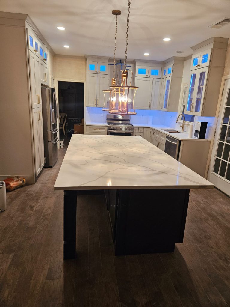 Kitchen with a white veined stone island on a dark painted base under two brass lantern pendants, white shaker cabinets with lit glass uppers