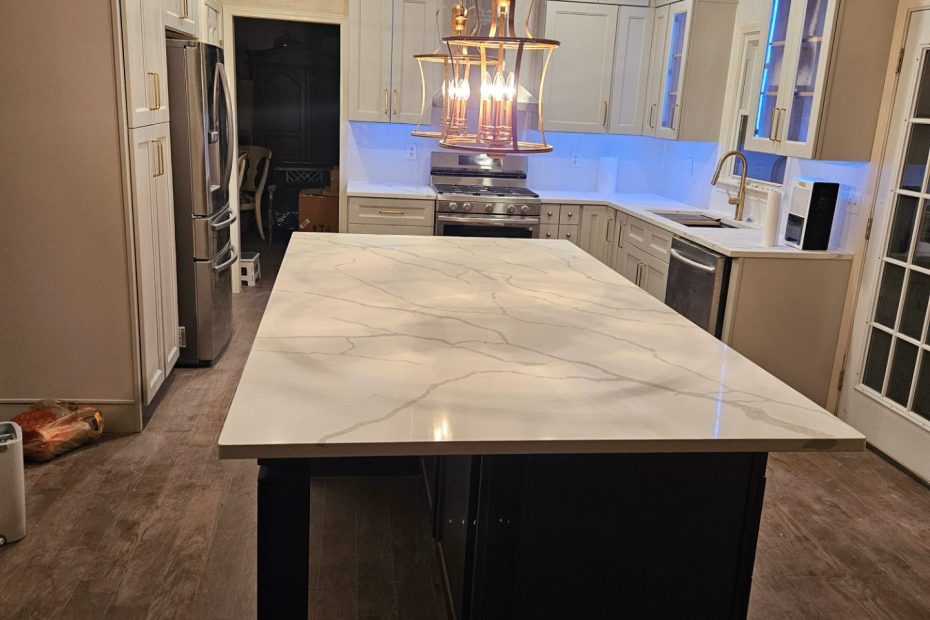 Kitchen with a white veined stone island on a dark painted base under two brass lantern pendants, white shaker cabinets with lit glass uppers