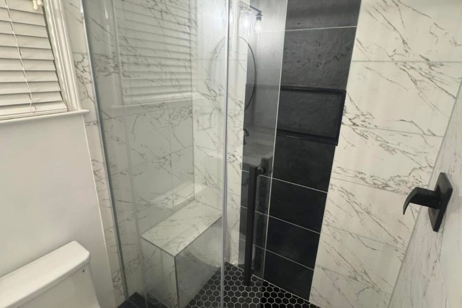 Walk-in shower with a frameless sliding glass door, white marble-look wall tile, a black accent stripe, a built-in bench and a black hexagon mosaic floor