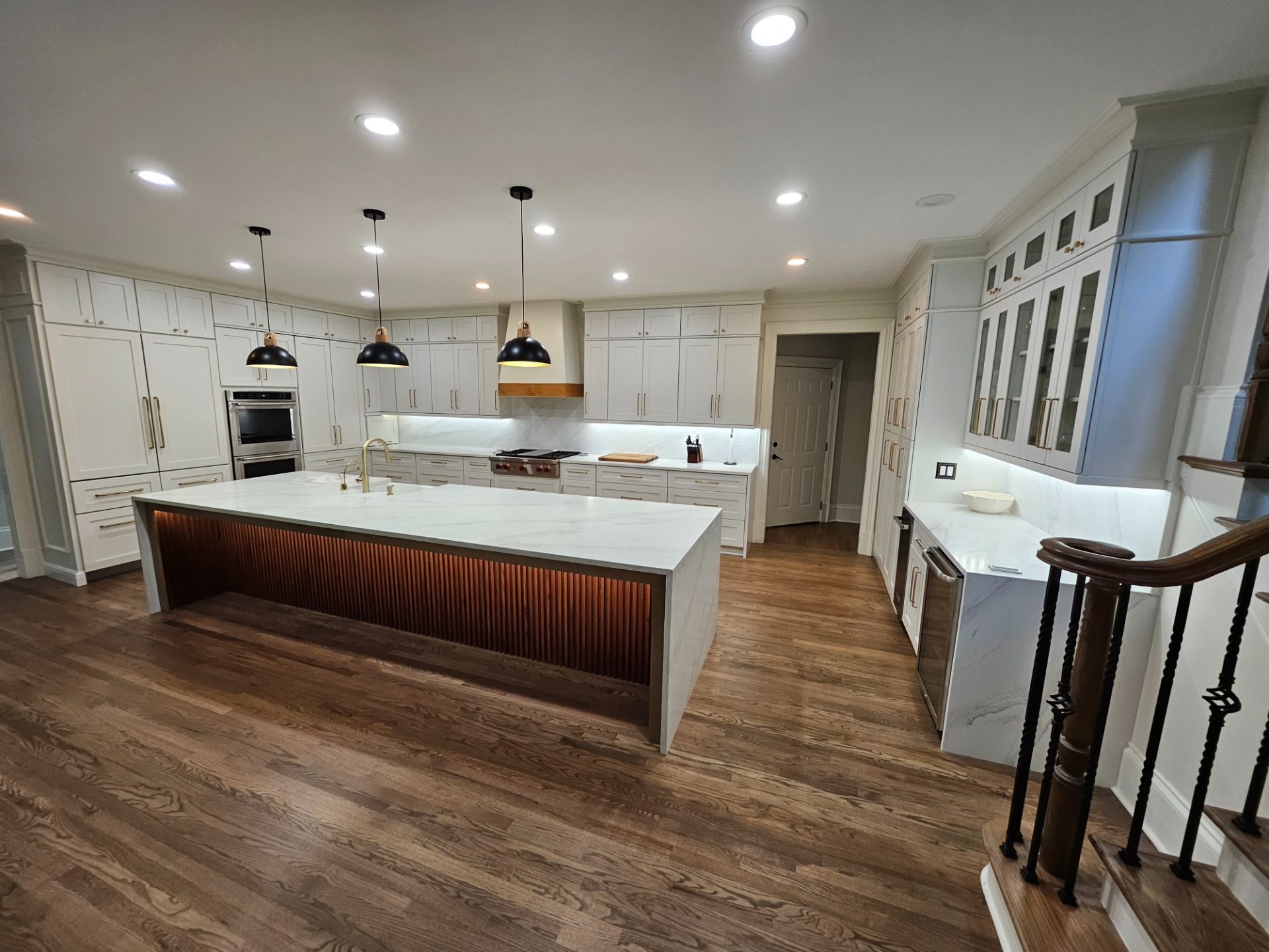 White kitchen with a long waterfall island over a lit wood-slat base, double wall ovens and glass-front uppers
