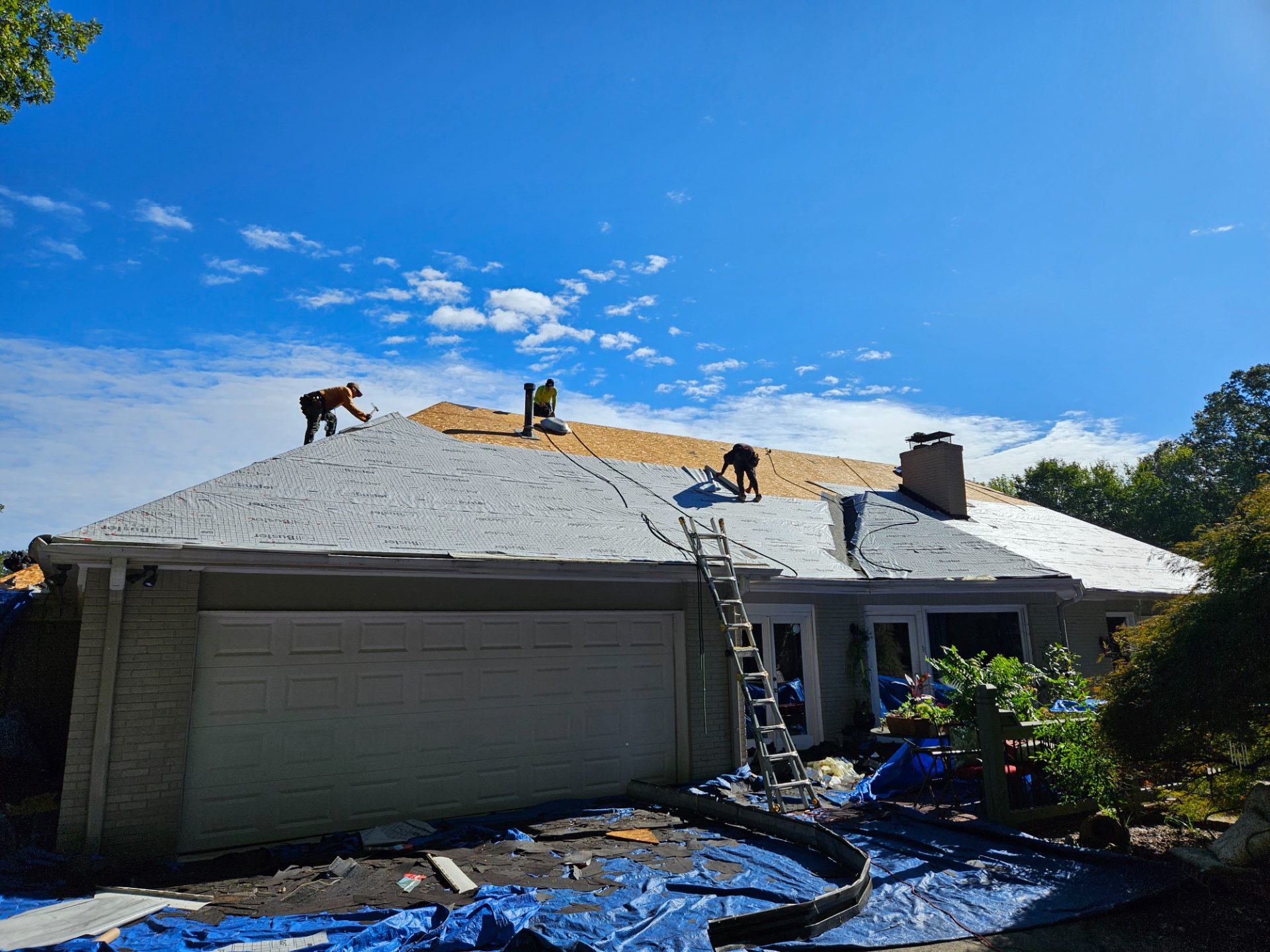 Crew laying gray synthetic underlayment over a torn-off brick ranch roof, with tarps covering the yard